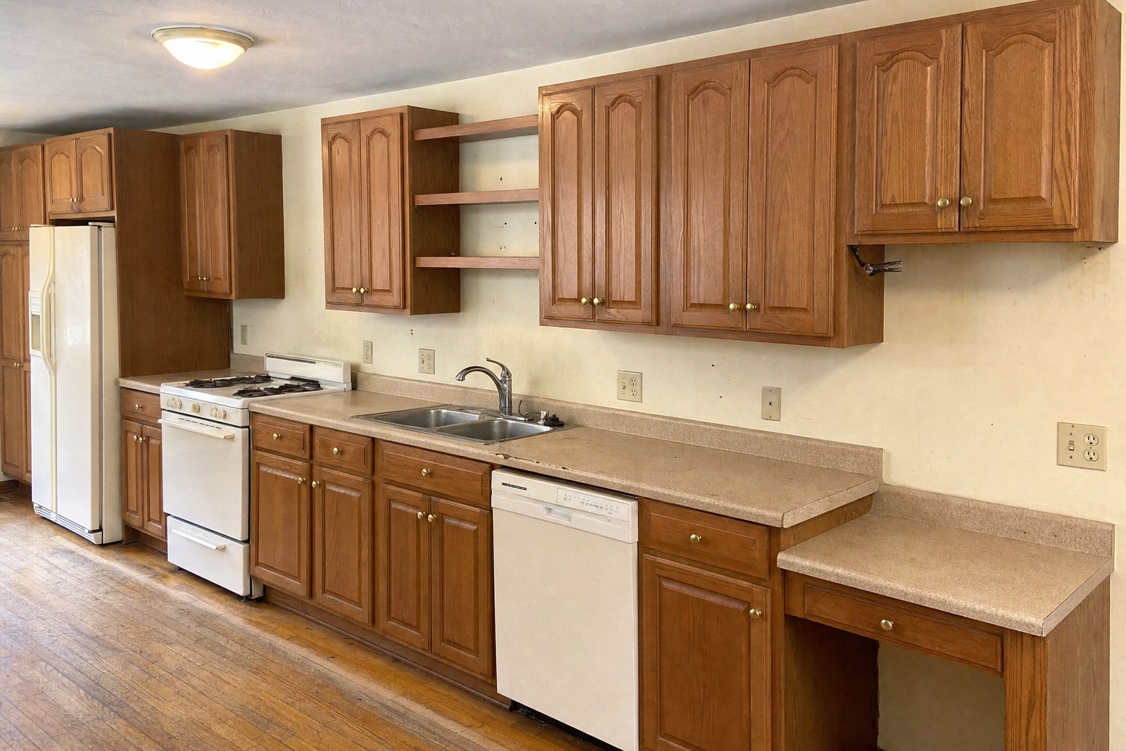 A dated 1990s kitchen — honey oak cabinets, worn laminate counters, no tile backsplash and yellowed white appliances