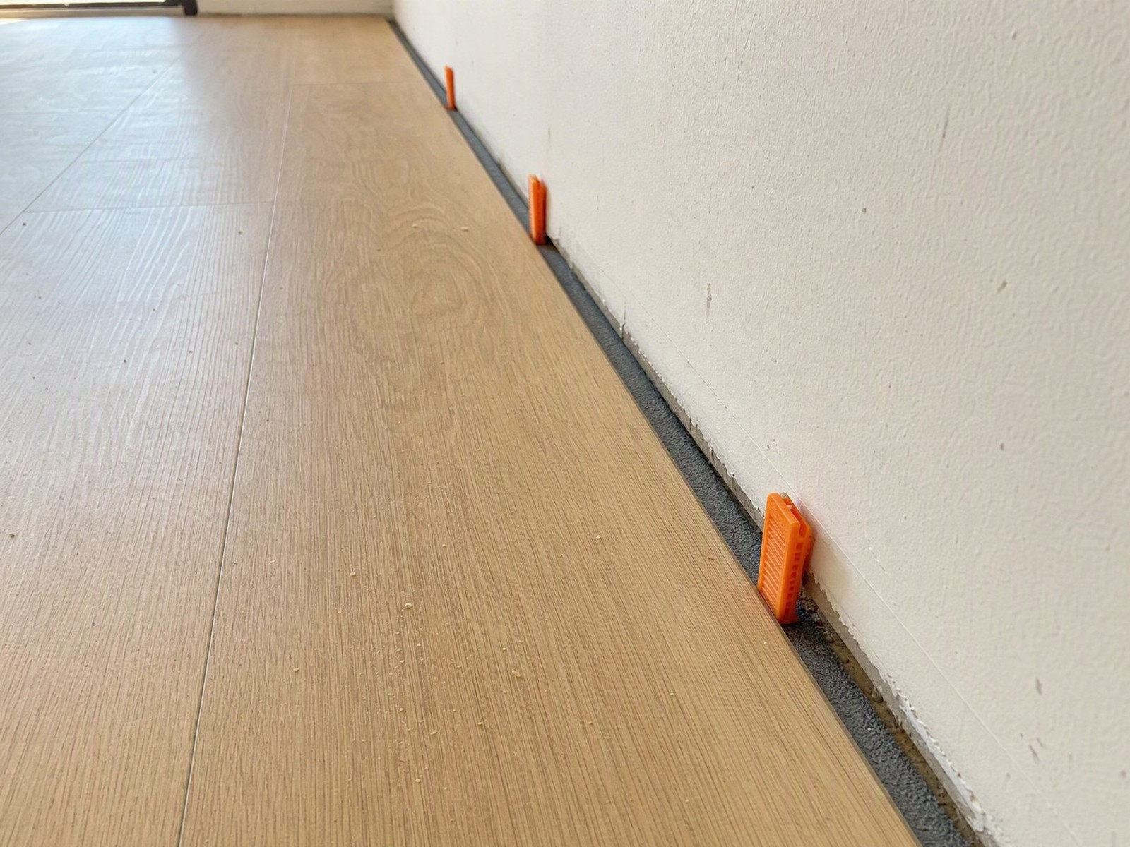 Orange spacer wedges holding a floating plank floor off the wall, leaving an even expansion gap before the baseboard goes on